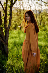 beautiful, a woman stands in a long orange dress, in the countryside, near a flowering tree, during sunset, illuminated from behind and looks sweetly into the camera