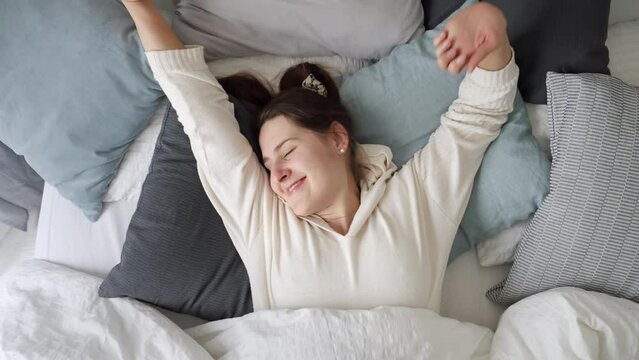 Relaxed young woman lying on soft pillows and stretching in bed at morning. Concept of comfort, relaxation, healthy sleeping and good start of new day