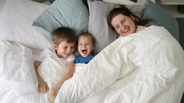 Happy laughing family with two kids hiding under blanket in bed and throwing it off. Concept of family happiness, relaxing at home, having fun in bed, parent and cheerful kids.