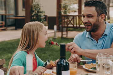 Cheerful father feeding his daughter with salad while having dinner outdoors together