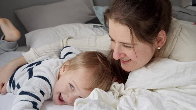 Young loving mother embracing her cute toddler son lying in bed at morning. Concept of family happiness, relaxing at home, having fun in bed, parent and cheerful kids.