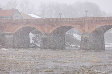 An old bridge from 1874 in Latvia made from bricks across a river on a cold and snowy Spring day
