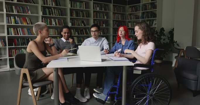 Pretty teenage student girl seated in wheelchair engaged in study, joint task preparation with diverse schoolmates gathered in library. Accessibility of education of person with physical disability