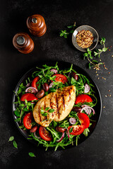 Chicken breast fillet grilled and fresh vegetable green salad with arugula, tomatoes and olives on black background, healthy food, mediterranean diet, top view - 595497244