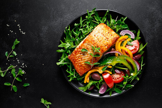 Salmon fillet grilled and fresh vegetable green salad of arugula with tomatoes, olives and bell pepper on black background, healthy food, mediterranean diet, top view