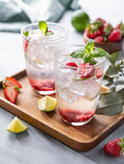 Fresh strawberry mojito in a glass with berries, lime, mint and ice in a wooden plate  on a light background. The concept of a summer refreshing drink for a party