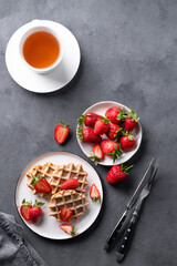 Homemade soft waffles with fresh strawberries and powdered sugar in a plate on a dark background with a cup of tea. Traditional Belgian waffles. Healthy breakfast.