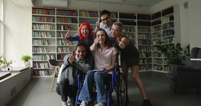 Pretty student girl with physical disability sits in wheelchair look at camera pose in library with schoolmates feel happy showing thumb up sign of success in study and friendship. Equality, education