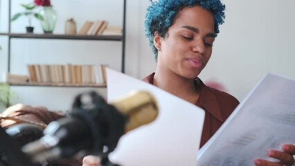 Young successful pretty African American woman journalist leads radio broadcast reading and discussing current news with listeners from sheets of paper sits in recording studio with microphone.