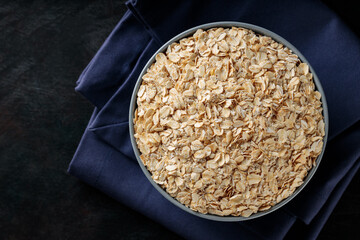 Raw oatmeal in bowl on a blue linen napkin. Oat flakes on a dark background. Healthy eating...