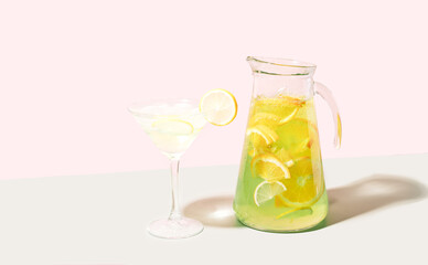 Glass goblet and decanter with drink with ice and lemon against pink background.