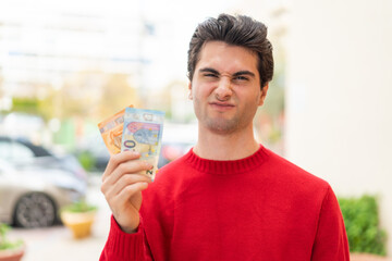 Young handsome man taking a lot of money at outdoors with sad expression