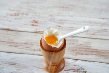 Soft-boiled egg in wooden egg cup showing runny yellow yolk with matching white ceramic spoon.