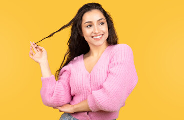 Cheerful millennial arab lady in casual holding hair, flirting, looking at empty space isolated on yellow background