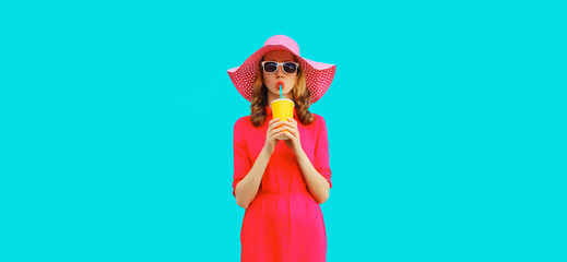Summer portrait of beautiful young woman drinking fresh juice wearing straw round hat, pink dress on blue background