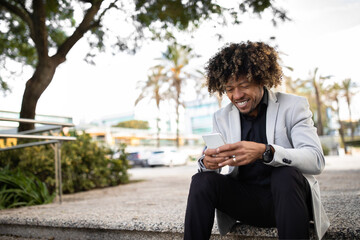 Successful black middle aged male entrepreneur using cellphone, texting or browsing internet, sitting on stairs outdoors