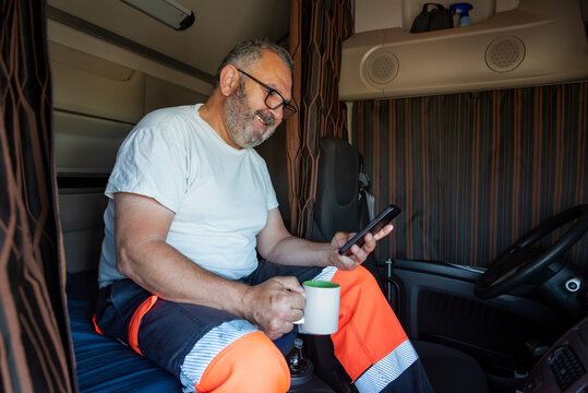 Senior Trucker Sitting In The Bed Of The Truck With The Curtains Drawn With A Cheerful Gesture Checking The Phone And A Cup Of Coffee In His Hand.