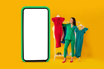 Shopaholic woman shopping online, standing near huge smartphone, choosing clothes over yellow background