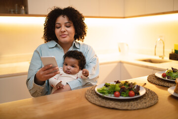 Young woman mother holding baby, have lunch, using smartphone