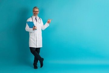 Smiling grey-haired mature caucasian doctor pointing at copy space - obrazy, fototapety, plakaty
