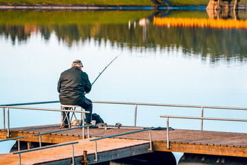 Senior man sitting at the lake jetty with rods and fishing equipment in the morning