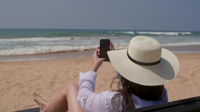 Woman tourist in sun hat taking picture of ocean on smartphone, back view. Brunette female sitting in sunbed on sandy beach resting on tropical resort. Tourism travel, vacation photographing moments.
