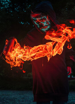 Masked figure with neon-lit mask and black hoodie, hands engulfed in flames. A captivating image that exudes intensity and energy.