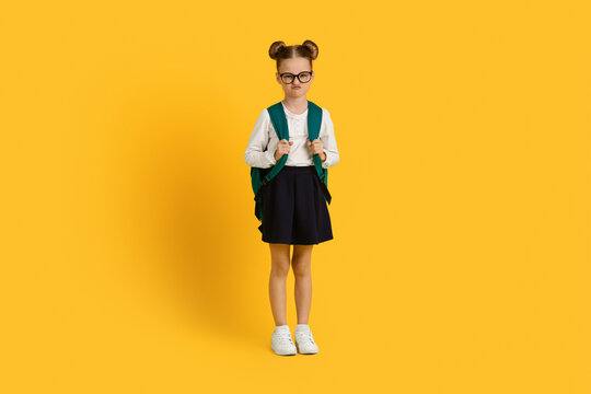 Portrait Of Grumpy Little Schoolgirl With Backpack Standing Over Yellow Studio Background