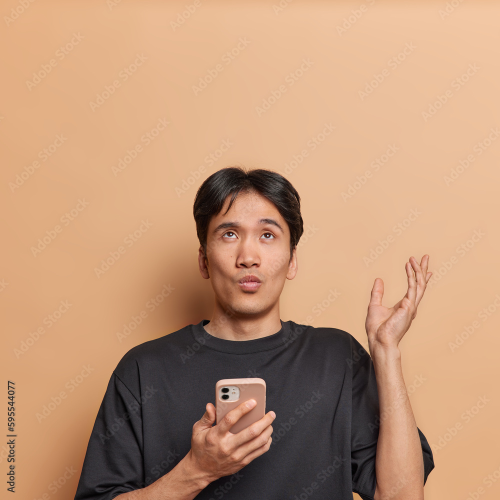 Wall mural Photo of puzzled uncertain Asian man poses with smartphone keeps palm raised up concentrated above dressed in casual black t shirt doesnt know why internet not working isolated over brown background - Wall murals