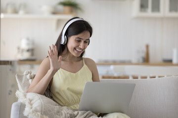 Cheerful pretty Hispanic young adult girl in big headphones enjoying online conversation, waving greeting hand at laptop webcam, smiling, laughing, enjoying Internet technology, connection