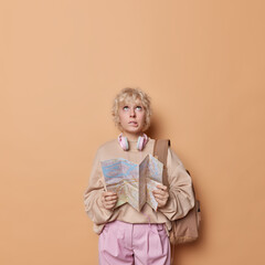 Puzzled female tourist holds travel city map thinks what to explore first bites lips and focused overhead carries rucksack on shoulder dressed casually isolated on brown background copy space for text