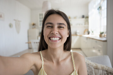 Cheerful excited pretty young adult girl taking selfie at home, holding gadget in outstretched hand, looking at camera with toothy smile, laughing, recording self video, talking on online call