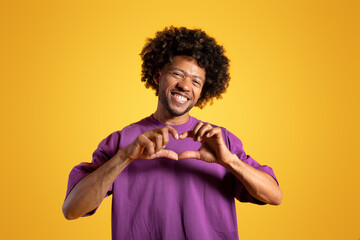 Happy funny adult african american curly man in purple t-shirt making heart gesture with hands