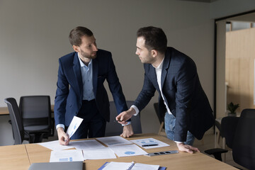 Two business men, colleagues, executives standing at meeting table with document, talking, discussing sales marketing report, project plan, brainstorming on statistic data for startup