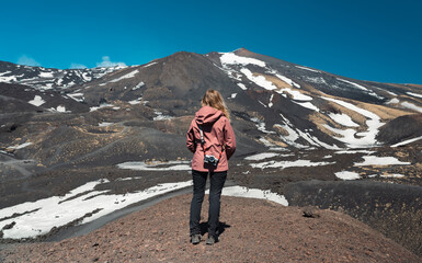 Obraz premium Blond photographer observes the panorama of southern slope of the Etna volcano. Back view of unrecognizable hiker with shoulder camera getting inspired to take a landscape photo. Adventure and outdoor