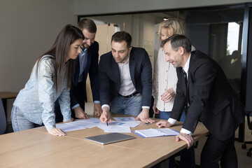 Diverse business team brainstorming on paper marketing reports, standing at meeting table with document, listening to project leader. Manager presenting startup statistic data to colleagues