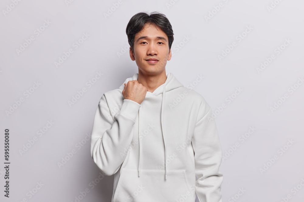 Wall mural Handsome dark haired Vietnamese guy looks directly at camera poses in studio for making photo dressed in casual hoodie isolated over white background ready for going out with friends during day off - Wall murals