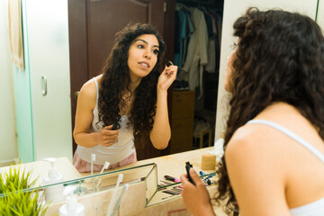 Hispanic woman looking in the bathroom mirror putting on makeup