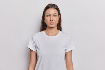 Waist up shot of nice looking woman has dark hair dressed in basic t shirt keeps arms down looks directly at camera isolated on white background. Brunette female model poses in studio for making photo - 595550262