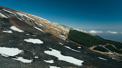 Snow melts on Etna volcano in spring. Panorama of uneven ground full of lava earth and craters. Small shrubs and large furrows, mediterranean climate. Colorful mountain trails on southern slope.