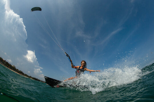 Kitesurfing girl in black sexy swimsuit with kite in sky on board in blue sea riding waves with water splash. Recreational activity, water sports, action, hobby and fun in summer time. 