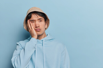 Bored Asian man holds hand on cheek looks tired aside stands in thoughtful pose concentrated aside...