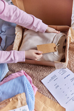 Cropped image of unknown woman holds tag with written price on folded jumper prepares pack product box puts invoice paper sells clothes online. Delivery preparation and online ecommerce concept