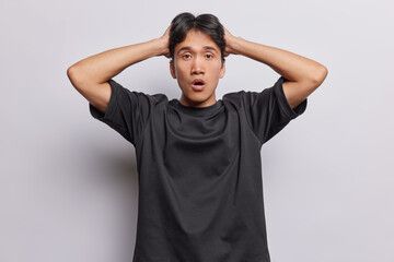 Waist up shot of emotional Chinese man with dark hair hears unexpected shocking news keeps hands on...