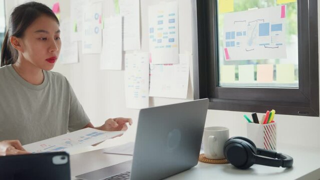 Young Asia girl IT development programmer typing on keyboard coding programming fixing data code on laptop computer screen follow code plan in tablet and paperwork on table workroom at house office.