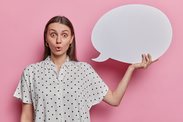 Surprised dark haired young woman dressed in polka dot blouse holds speech bubble poses with empty...