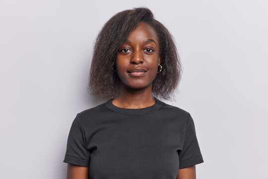 Portrait of lovely teenage girl with dark skin short afro hair poses against white background with both arms down dressed in casual black t shirt looks directly at camera. Neutral facial expression