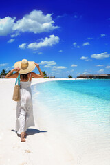 A elegant luxury woman in a white dress and hat walks down a tropical paradise beach in the Maldives islands