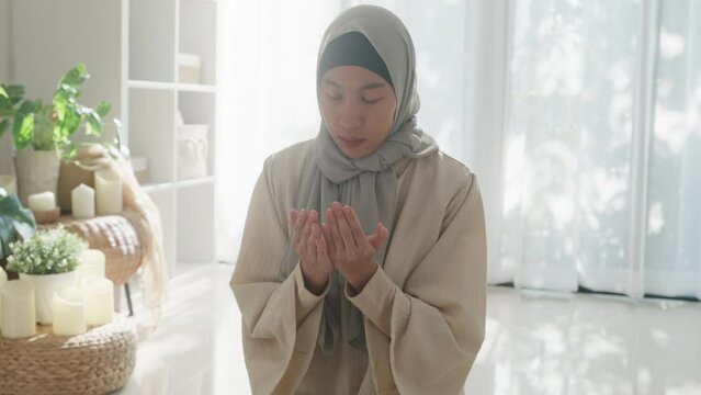 Asian muslim woman sitting on prayer mat pray to allah wear prayer clothes green mukena cover hijab in living room at home, praying room bright light, Islamic faith, Ramadan Kareem, Eid Mubarak.