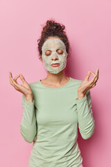 Relaxed dark haired woman applies moisturising soap mask on face keeps eyes closed meditates to relax undergoes beauty procedures wears casual jumper isolated over pink background. Beauty concept